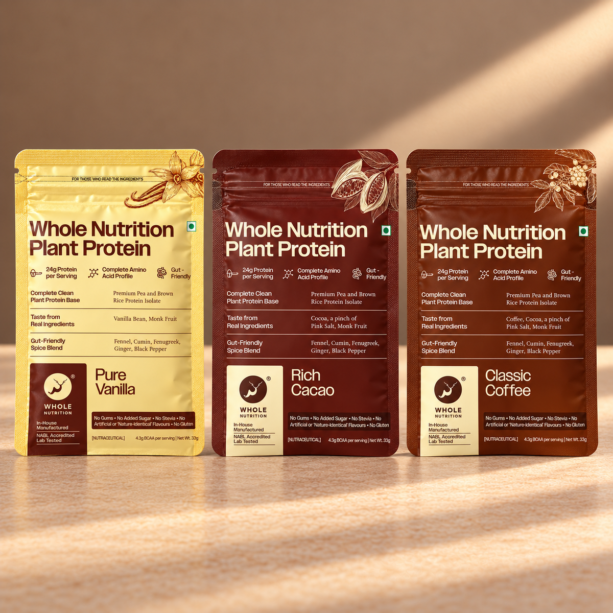 Three stand-up pouches of Whole Nutrition Plant Protein powder are arranged on a wooden surface under warm light. The left pouch is Pure Vanilla in a pale yellow color, and the middle and right pouches are Rich Cacao and Classic Coffee in dark brown.